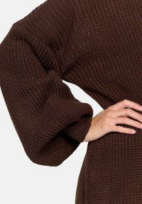 Brunstickad tröja med en texturerad mönster, löst sittande ärmar och ribbade muddarna. Tyget verkar mjukt och tjockt.
