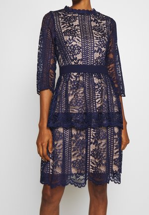 Robe en dentelle florale bleu marine avec bords festonnés, manches trois-quarts, taille cintrée et détail volanté en couches sur une jupe mi-longue.