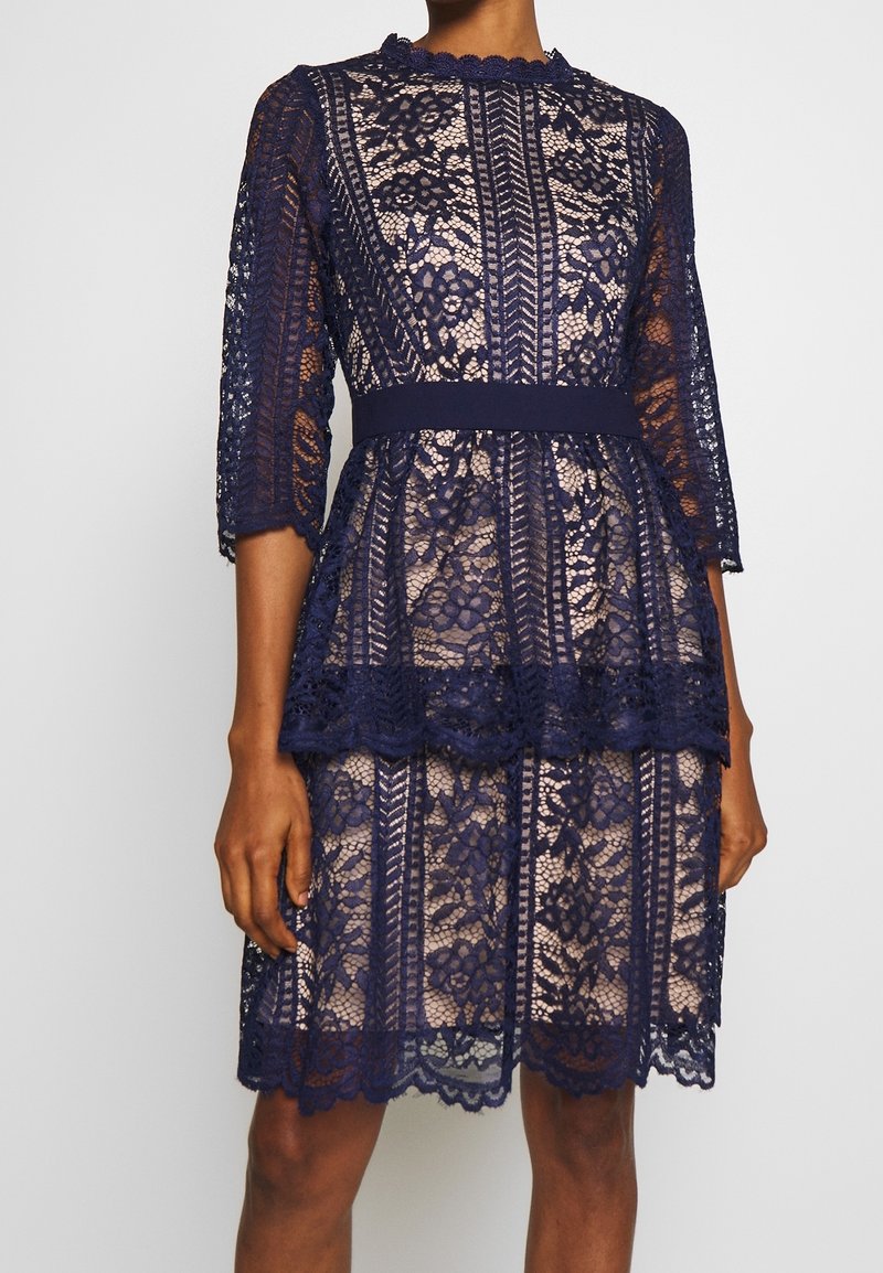 Robe en dentelle florale bleu marine avec bords festonnés, manches trois-quarts, taille cintrée et détail volanté en couches sur une jupe mi-longue.