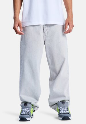 Mężczyzna ubrany w luźne, jasnoszare dżinsy, białą koszulkę i szare trampki z neonowymi zielonymi akcentami, stojący na białym tle.