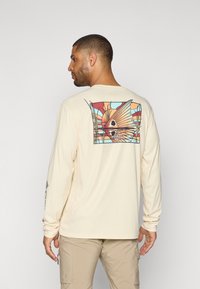Camicia a maniche lunghe color crema con una grafica colorata di un pesce sulla schiena. Presenta una vestibilità rilassata e polsini a costine.