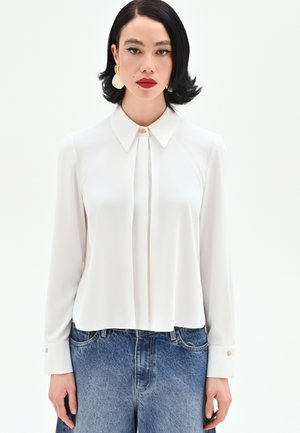 Donna con capelli neri corti che indossa una camicetta bianca a maniche lunghe con colletto e jeans blu, in piedi davanti a uno sfondo bianco uniforme.