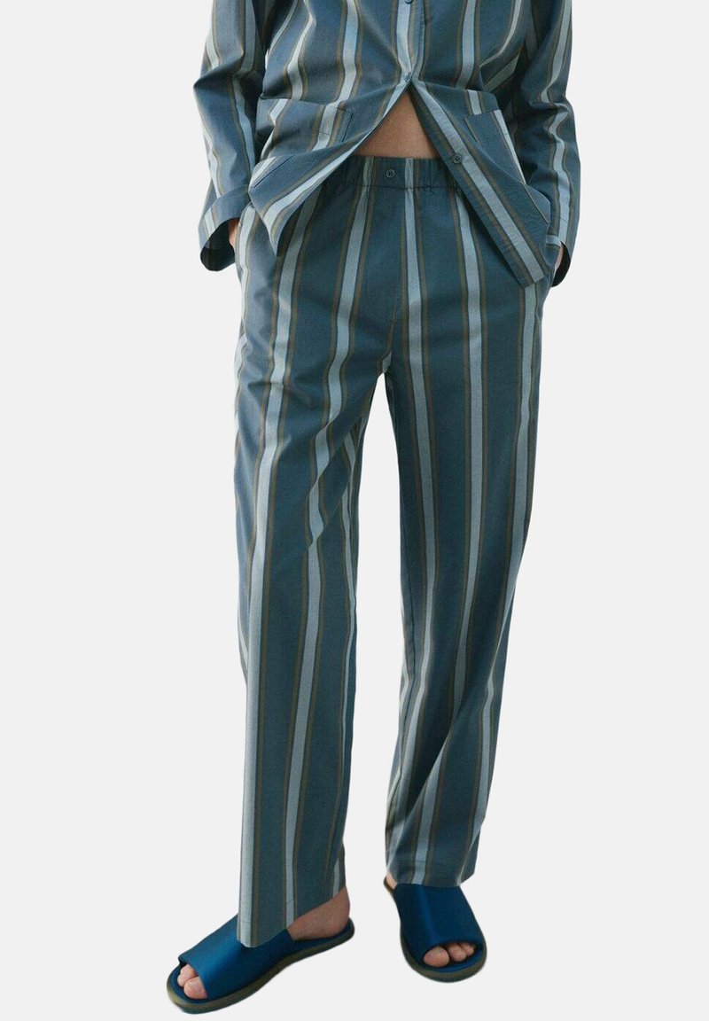 Osoba nosiaca voľné nohavice a košeľu so zvislými pásmi v modrej, sivej a béžovej farbe, doplnené tmavomodrými otvorenými sandálmi.