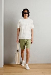 Hvid skjorte med korte ærmer og striber, grønne shorts, hvide sneakers, sorte solbriller og en beige tote bag. Enkel design og letvægtigt stof.