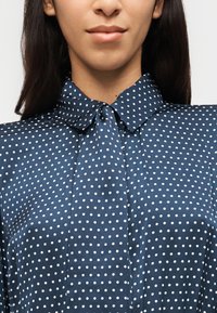 Femme portant un chemisier bleu marine à pois blancs avec un col noué en forme de nœud, montrée de la hauteur des épaules aux lèvres sur un fond uni.