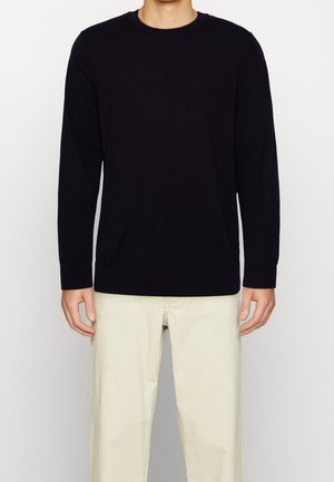 Homme portant un pull noir uni à manches longues et un pantalon écru, debout devant un fond neutre.