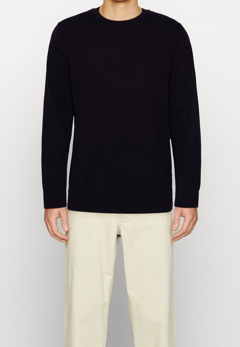 Homme portant un pull noir uni à manches longues et un pantalon écru, debout devant un fond neutre.