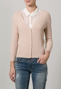 Cardigan rose pâle en maille côtelée avec des manches trois-quarts, fermeture par boutons et décolleté en V, porté par-dessus une chemise blanche à col.