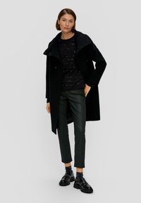 Czarny wełniany płaszcz z wysokim kołnierzem, noszony na czarnym swetrze w wzory i ciemnozielonych spodniach; błyszczące czarne buty na grubych podeszwach.