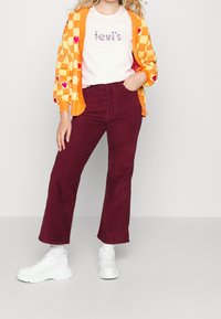 Orange checkerboard cardigan with red heart accents over a white t-shirt featuring "Levi's" in glitter, paired with burgundy corduroy pants and white sneakers.