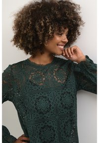 Blusa in pizzo verde scuro con intricati motivi floreali, maniche lunghe e scollatura a onda, su uno sfondo neutro.