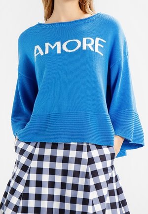 Mujer con un suéter azul con la palabra "AMORE" y una falda a cuadros en blanco y negro, con las manos en los bolsillos, sobre un fondo blanco.