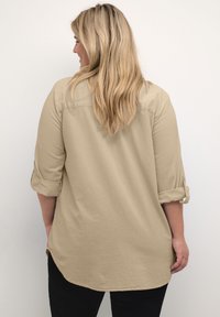 Chemise beige à boutons avec des manches retroussées, détail de pli au dos, ourlet arrondi et un tissu doux et texturé. Portée avec un pantalon noir.