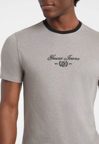 Sivá bavlnená tričko s krátkymi rukávmi, s čiernym okrúhlym výstrihom a vyšitým logom "Guess Jeans" v čiernej farbe na hrudi.