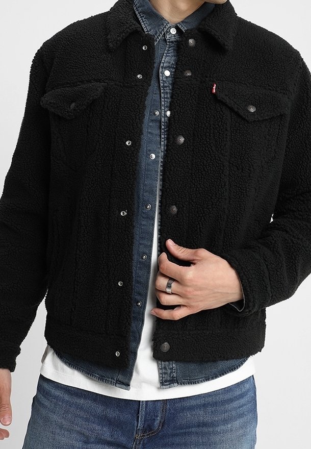 Homme portant une veste en sherpa polaire noire par-dessus une chemise en jean bleue et un t-shirt blanc, la main ajustant les boutons avant de la veste.