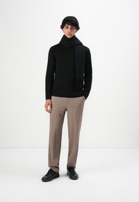 Jonge man staat, gekleed in een zwarte trui en sjaal, beige broek en zwarte sneakers, tegen een effen witte achtergrond.