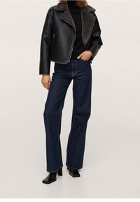 Giacca di pelle nera con colletto in shearling grigio, abbinata a jeans a campana blu scuro. Il modello indossa stivali neri con tacco. Sfondo semplice.