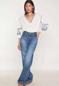 Blusa bianca ricamata con accenti floreali blu, abbinata a jeans blu a gamba larga con vita alta. Tessuto testurizzato, vestibilità rilassata.