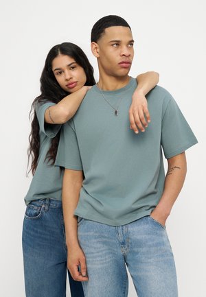 Δύο νεαροί ενήλικες φοράνε ανοιχτά γαλάζια-πράσινα πουκάμισα και τζιν, στέκονται κοντά, με τη γυναίκα να ακουμπάει το χέρι της στον ώμο του άντρα.