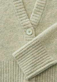 Cardigan en maille beige doux avec col et poignets côtelés, doté d'un grand bouton rond unique à l'avant.