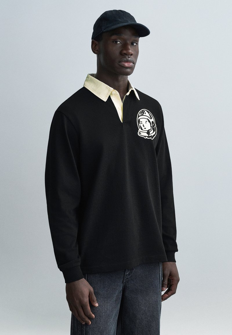 Μαύρο πόλο με μακριά μανίκια και κίτρινο πουκάμισο με γιακά από κάτω. Διαθέτει καρτούν γραφικό με αστροναύτη στη αριστερή πλευρά. Φορεμένο με σκούρο τζιν.