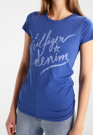 Donna che indossa una maglietta aderente blu con "Hilfiger denim" scritto in corsivo e jeans azzurri chiari su uno sfondo bianco semplice.