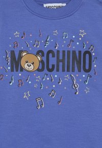 Blå skjorta med svart "MOSCHINO" text, ett björns ansikte som ersätter "O," omgiven av färgglada musiknoter och stjärnor.