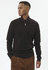 Man wearing a dark brown quarter-zip sweater and brown corduroy pants, looking to his left with hands in pockets against white background.