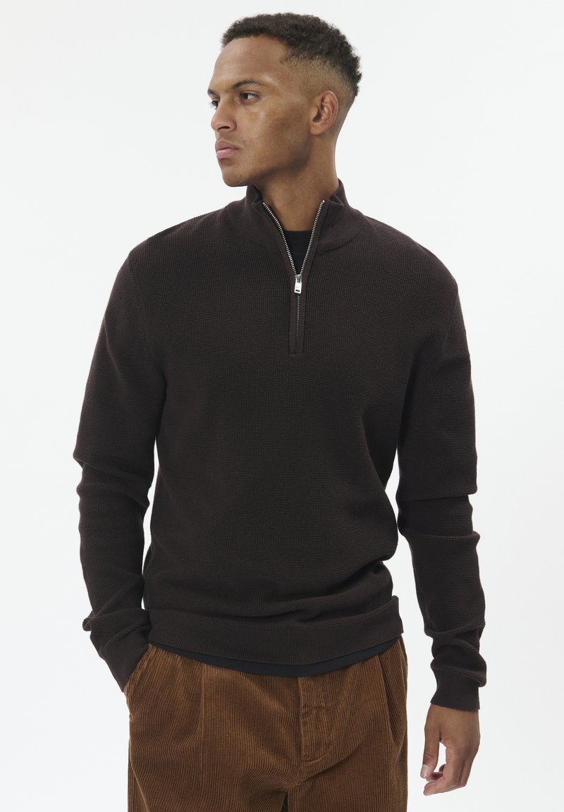 Man wearing a dark brown quarter-zip sweater and brown corduroy pants, looking to his left with hands in pockets against white background.