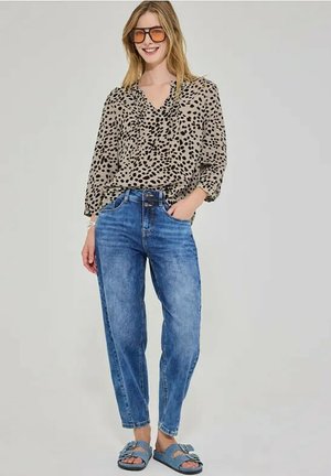 Kvinde iført leopardprint bluse, blå højtaljede jeans, blå sandaler med spænder og store tonede briller, smilende med den ene hånd i lommen.