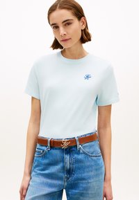 T-shirt en coton bleu clair à manches courtes et col rond, orné d'un logo brodé bleu sur la poitrine. Porté avec un jean en denim taille haute.