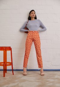 Calças laranja com padrão de leopardo e cintura alta, combinadas com um suéter acolchoado azul claro. Uma bolsa castanha e um banco de acrílico transparente estão por perto.