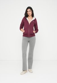 Μπορντό φούτερ με φερμουάρ μπροστά και τσέπες, συνοδευόμενο από γκρι φόρμα. Το φούτερ διαθέτει λευκό γιακά με επένδυση από φλις και λεπτή εφαρμογή.