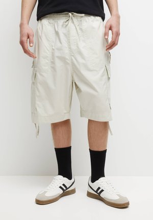 Mann trägt beige Cargoshorts mit Kordelzugbund, schwarze Socken und weiße Turnschuhe mit schwarzen Streifen vor einfarbigem Hintergrund.