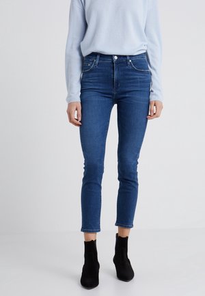 Kvinde iført blå skinny jeans med høj talje og sorte ankelstøvler, kombineret med en lyseblå langærmet top, står på et hvidt gulv.