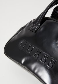 Schwarze Handtasche aus patentiertem Leder mit geprägt "GUESS"-Logo, runder Form und doppelten Tragegriffen. Sie zeichnet sich durch eine glänzende Oberfläche und genähte Akzente aus.