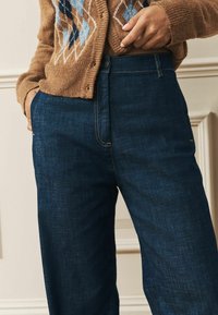 Tumši zili džinsu platleju bikses ar gludu tekstūru un redzamām šuvēm. Kombinētas ar brūnu, rakstainu adītu kardigānu.