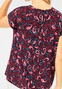 Kortärmad blus med en mörk marinblå botten och mönster av rosa och vita paisleymönster, prydd med små blå blomdetaljer och en nyckelhålsrygg.