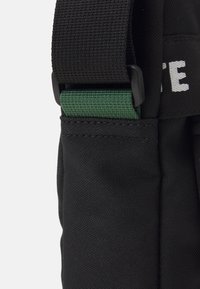 Black nylon bag featuring a textured finish, adjustable strap with green accents, and printed white lettering on the side.
