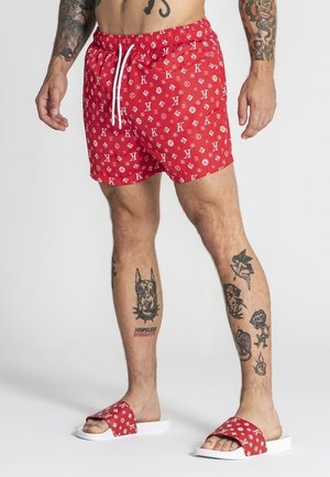 Mężczyzna w czerwonych kąpielowych szortach z wzorem i pasujących klapkach, pokazujący tatuaże na ramionach i nogach na jednolitym jasnym tle.
