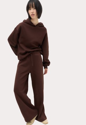 Kvinde med kort mørkt hår iført en løs brun hættetrøje og bukser med vide ben, der står med den ene hånd bag ryggen og beige sko.
