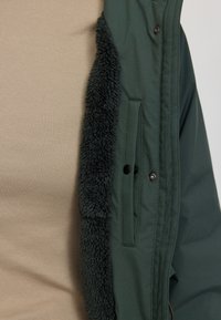 Grüne Jacke mit Fleece-Futter entlang des Reißverschlusses, mit einer Seitentasche und schwarzen Druckknöpfen. Darunter befindet sich ein beigefarbenes gestreiftes Shirt.