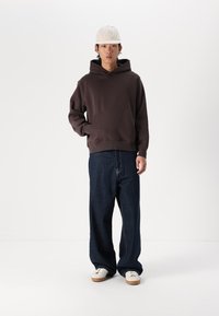 Brown hoodie with a front pocket, paired with loose-fitting dark blue jeans and white sneakers. Model wears a light-colored cap.