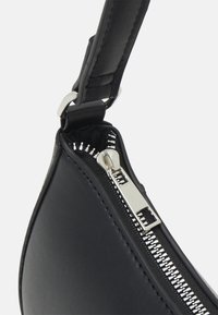 Nærbillede af hjørnet på en sort lædertaske med sølvfarvet metallynlås og syet stropfastgørelse.