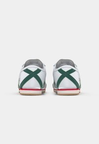 Zapatillas blancas con rayas cruzadas verdes y suelas rojas, vistas desde atrás sobre un fondo blanco plano.