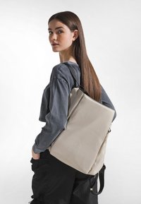 Mochila beige con una forma rectangular elegante, hecha de un material liso, con un logo en relieve en la parte delantera y correas ajustables.