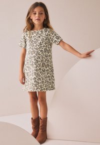 Jeune fille en robe à imprimé léopard et bottes marron, debout avec une main posée sur une surface blanche incurvée.