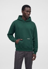 Homme portant un sweat à capuche vert foncé avec une poche avant et un pantalon gris, debout devant un fond clair uni.