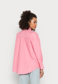 Camisa de algodão rosa com um corte solto, mangas longas e uma bainha curva. Apresenta um discreto detalhe de logo nas costas. Usada com calças de ganga claras.