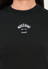 Čierne bavlnené tričko s okrúhlym výstrihom a bielym potlačeným textom: "MOSCHINO 86 · 23 JEANS," umiestnené na strede hrudníka.